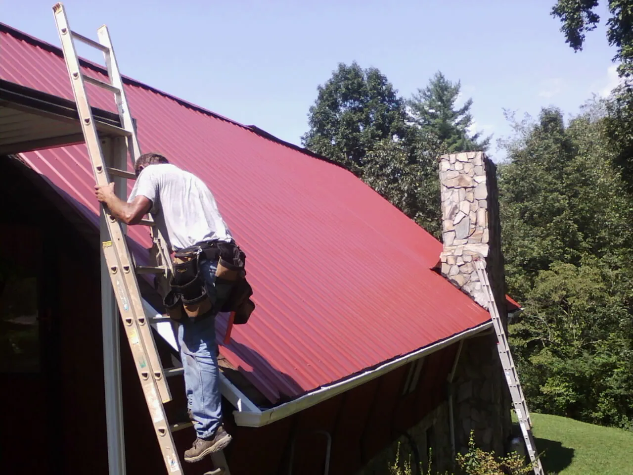 Roofer on a ladder beside a red metal roof and stone chimney — Soffit & Fascia Repair by Roofing Brothers in Turlock